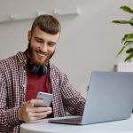 Happy young attractive ginger bearded man working at a laptop while sitting in a cafe, wearing in basic clothes, broadly smiling and looking at his smartphone.