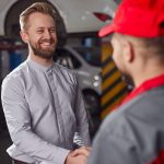 Content adult bearded male client shaking hand of unrecognizable repairman after getting car fixed in professional automobile repair service workshop